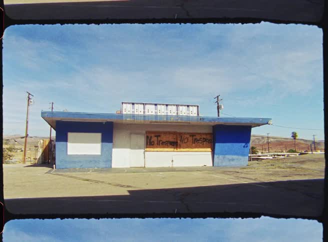 An abandoned singlestory blue building stands in a desert landscape with no trespassing signs spraypainted across boardedup windows the scene is captured with a vintage film border showing power lines and distant hills under a clear sky