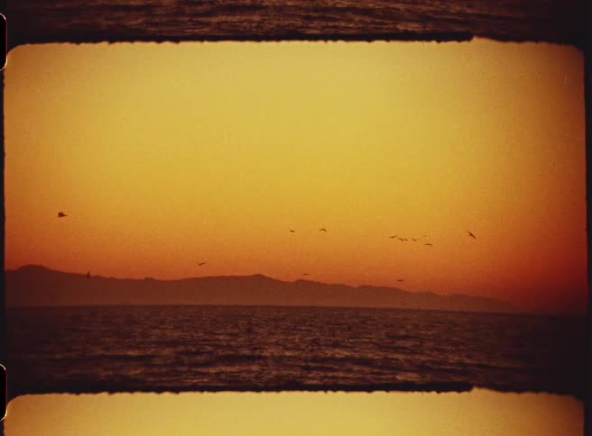 A flock of birds flies low across the horizon above a vast body of water during a warm sunsetthe scene is framed with a dark vintage film border and features a hazy mountain range in the distance