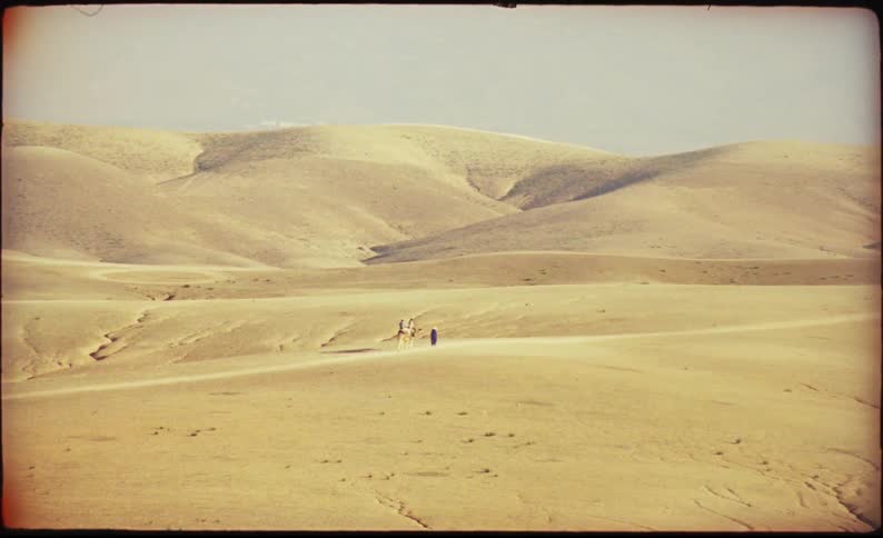 Tiny figures walk across vast rolling sand dunes under a hazy sky