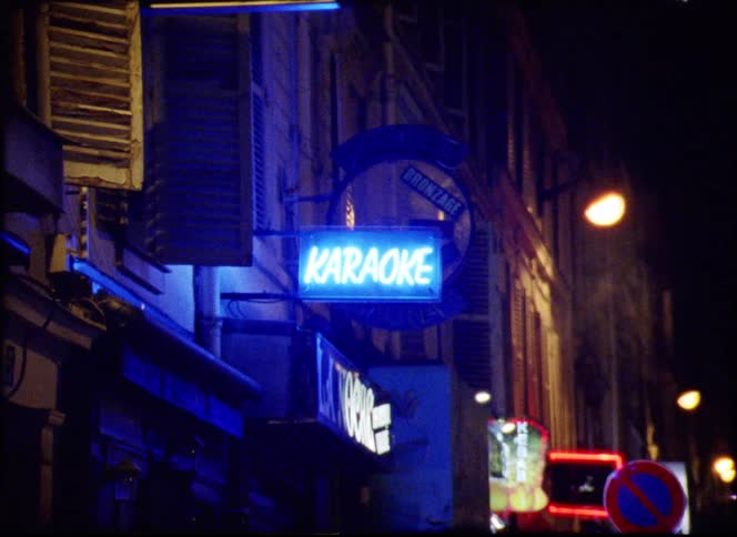 A glowing karaoke sign lights up a dim neon lit street corner at night