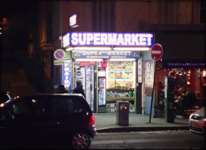 A supermarket on the side of the street at night its supermarket sign glowing above with a stop sign nearby as cars drive past