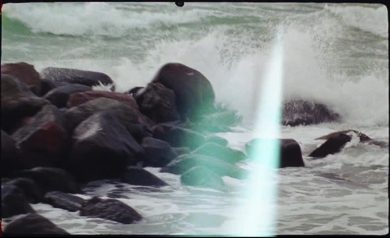 A powerful ocean wave crashes against dark rounded coastal rocks sending white spray and foam swirling around the slick stone surfaces under an overcast sky
