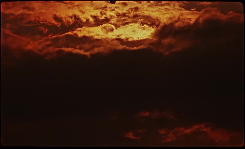 This video shows a dramatic sunset or sunrise with thick swirling clouds illuminated by a deep orange and golden glow the footage uses a vintage film style with a dark moody aesthetic and visible film grain