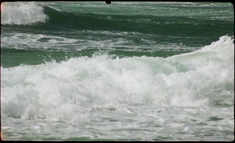 The video shows multiple layers of ocean waves crashing and turning into white foam the footage has a vintage grainy film aesthetic with visible frame borders and motion