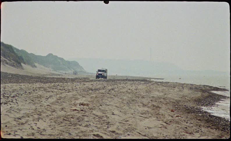 A dark offroad vehicle drives along a wide sandy beach between grassy dunes and the ocean tide under a hazy sky