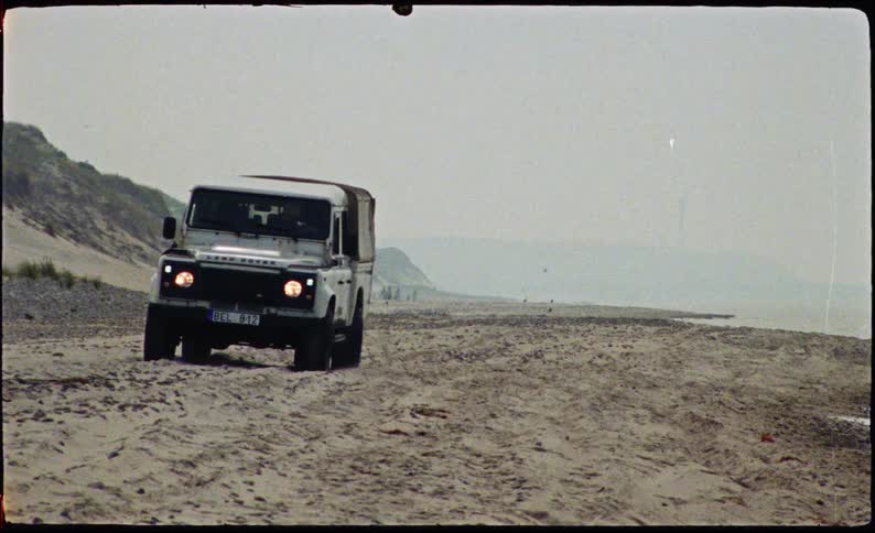 A white land rover drives across a wide sandy beach next to the ocean under an overcast sky while distant figures and a thin structure are visible on the hazy coastline