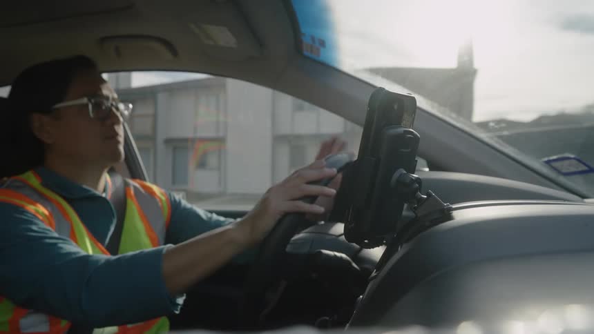 A parcel delivery driver in a safety vest drives through a city on a sunny day using a dash mounted phone for navigation