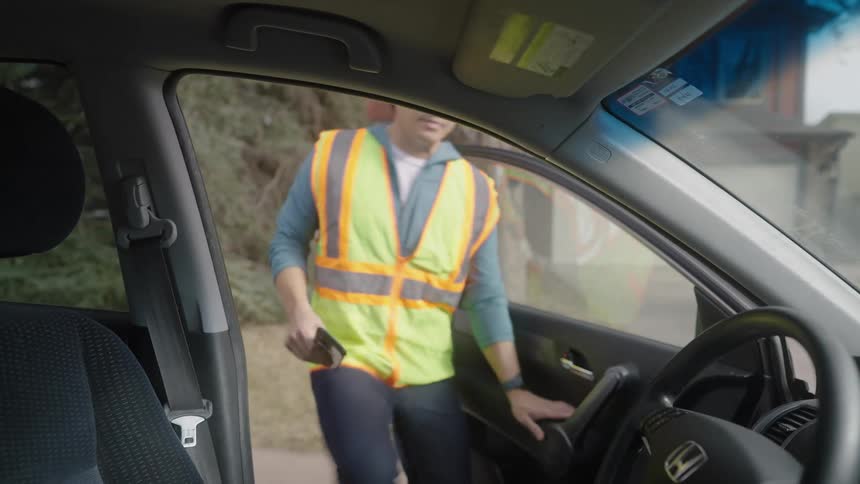 A man wearing a green vest rushing into a car at midday holding a smartphone on the side of the street