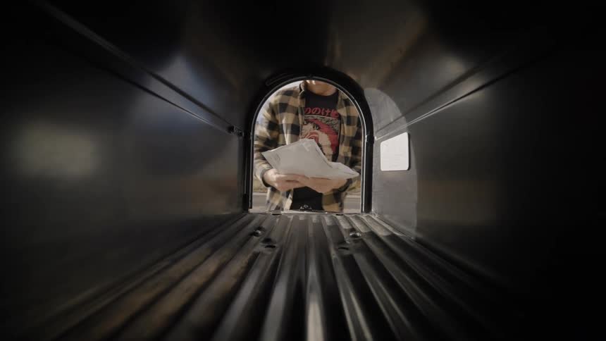 A man in a plaid shirt reaches into a dark mailbox to retrieve several packages and letters checking the contents before closing the door on a bright sunny day