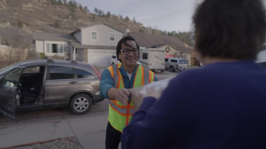 A delivery driver wearing a highvisibility safety vest smiles and hands a brown cardboard package to a person standing outside of a car in a residential neighborhood