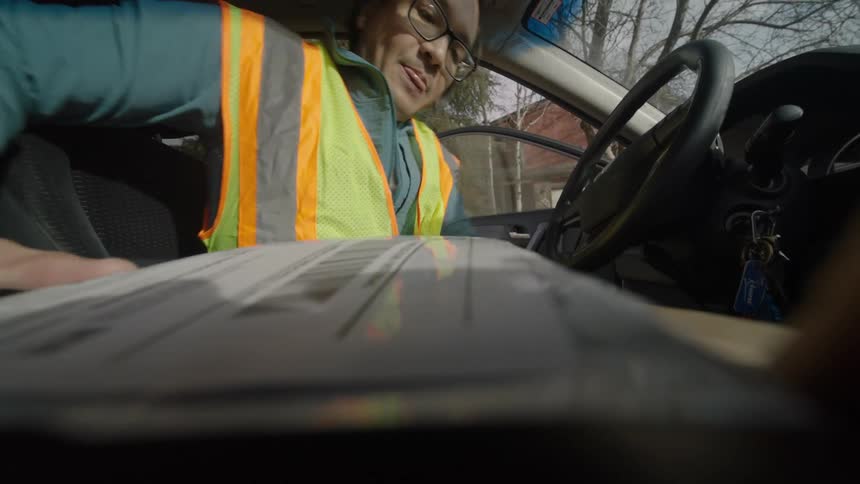 A delivery driver wearing a safety vest sits in a car scans a handheld package with a device and then opens the driverside door to exit the vehicle