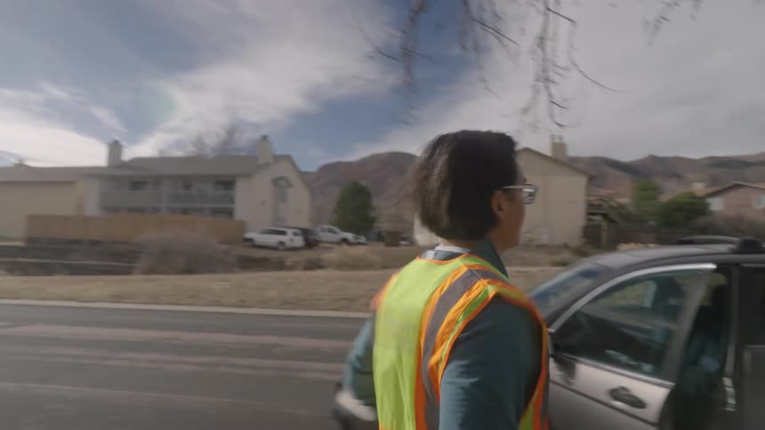 A man wearing a highvisibility safety vest jogs down a quiet suburban sidewalk before reaching a parked car opening the door and preparing to climb into the drivers seat under a bright clear sky