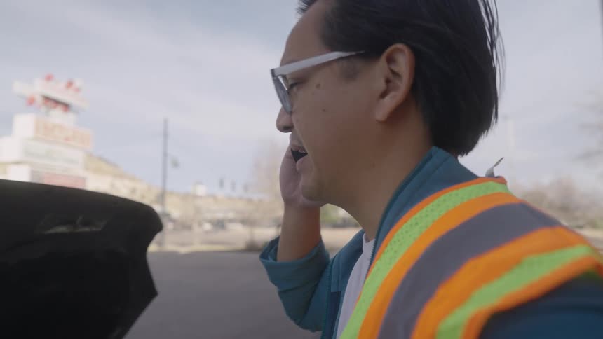 A construction worker wearing a green vest and glasses standing outside a building at midday making a phone call