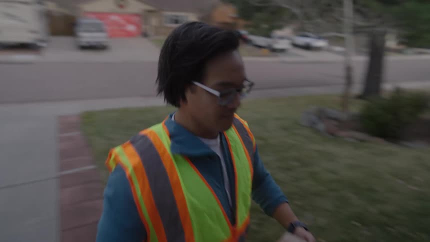 A delivery worker in a safety vest walks up a residential driveway carrying a cardboard package they reach the front door and place the parcel carefully on the concrete porch before walking away