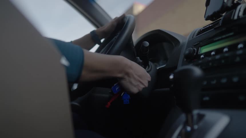A driver reaches toward the ignition and turns the key to start a vehicle while holding onto the steering wheel the camera angle captures a closeup of the dashboard and center console from the passenger side
