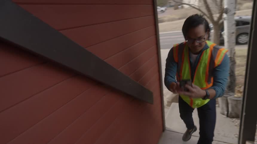A delivery worker wearing a safety vest places a cardboard package on a residential doorstep then uses a smartphone to take a confirmation photo of the item before walking away down the wooden stairs toward the street