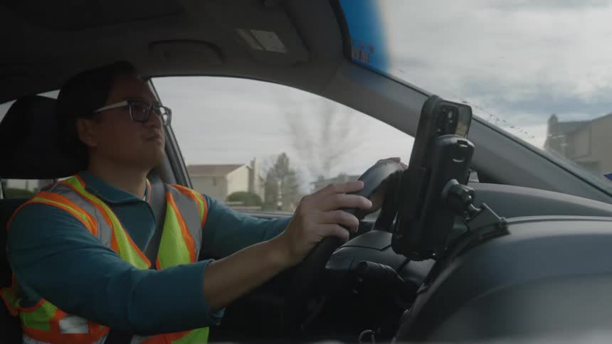 A person wearing glasses and a highvisibility safety vest is driving a car through a residential area they briefly adjust a smartphone mounted on the dashboard before taking it out of the holder to make a phone call while continuing to drive