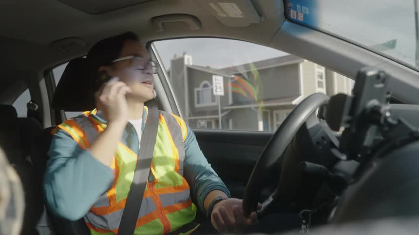 A man wearing a green vest driving down the street at midday talking on the phone