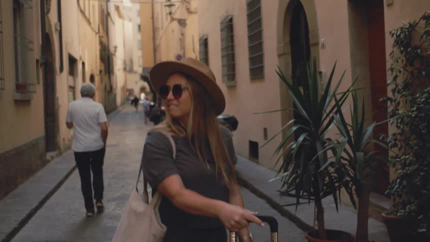 Woman wearing a hat and sunglasses carrying a bag walks through an urban area
