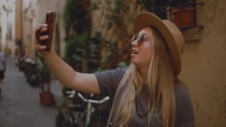 Woman uses a smartphone to take a selfie on a narrow street with old buildings and potted plants