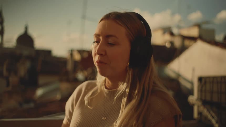 Woman wears headphones while talking on a balcony overlooking the city at sunset