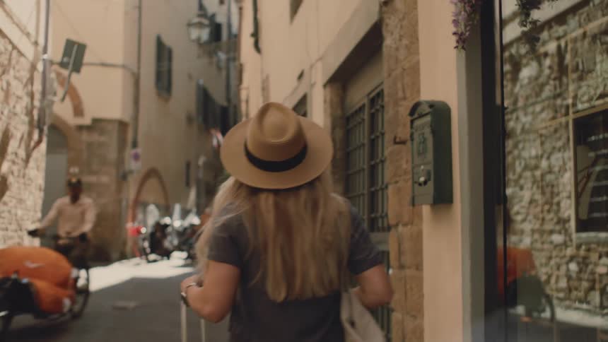 Woman wearing a hat and carrying a suitcase walks down an urban corridor