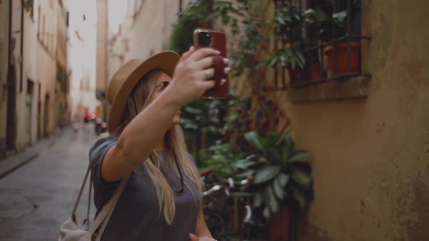 A young woman in an alleyway at midday holding a handbag and a suitcase wearing a hat and sunglasses her long blonde hair flowing as she records a selfie video on her phone