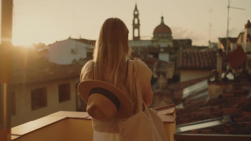 A woman with long blonde hair stands on a balcony and takes a photo of a european cityscape during sunset she wears a widebrimmed hat hanging on her back as she looks out toward a large dome and clock tower in the distance