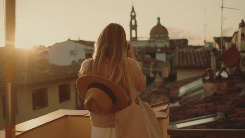 A young woman sitting on a rooftop at sunset her hat resting behind her as she photographs the surrounding environment