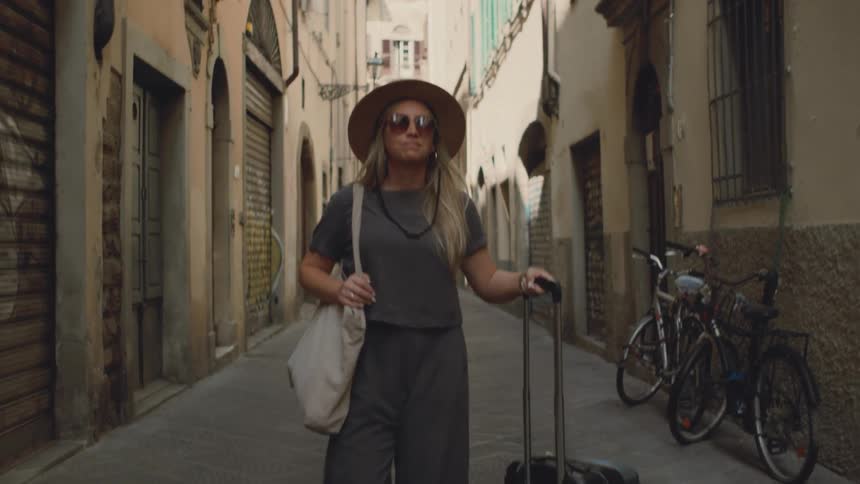 Woman walks down a narrow street pulling a suitcase and carrying a shoulder bag