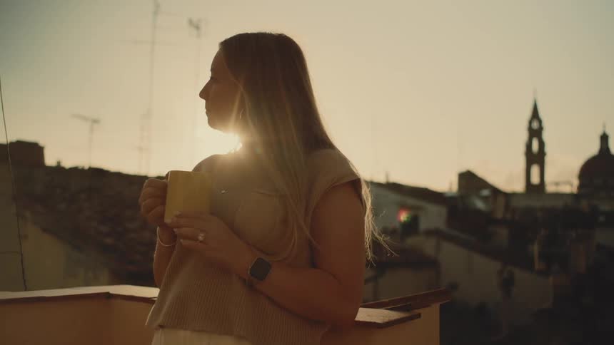 Woman holds a mug on a balcony while looking over the urban landscape at sunset
