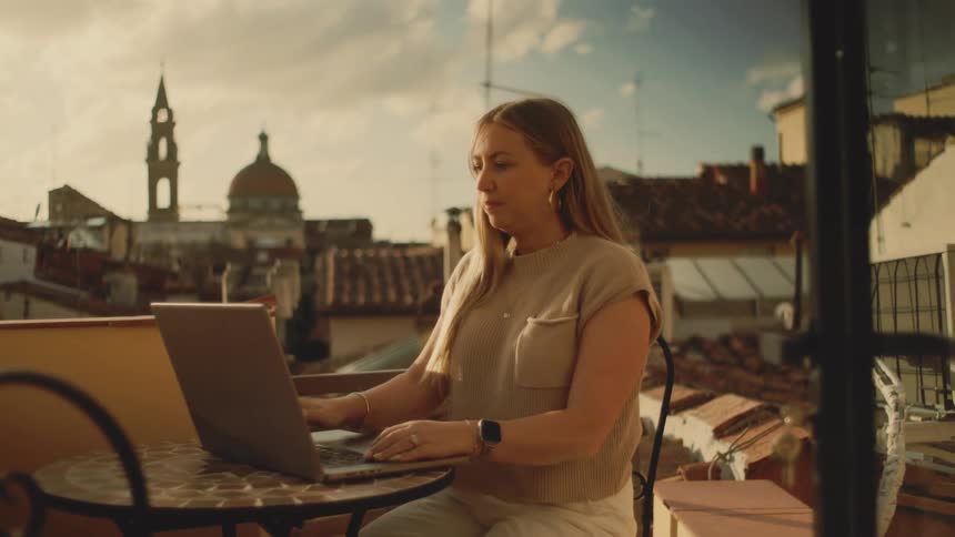Woman sitting at an outdoor table typing on a laptop computer as she stops to take a drink from her cup