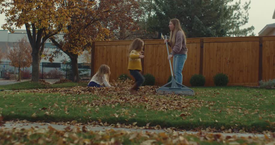 Two young girls playing in the front yard and throwing leaves into the air as a teenage girl rakes the leaves
