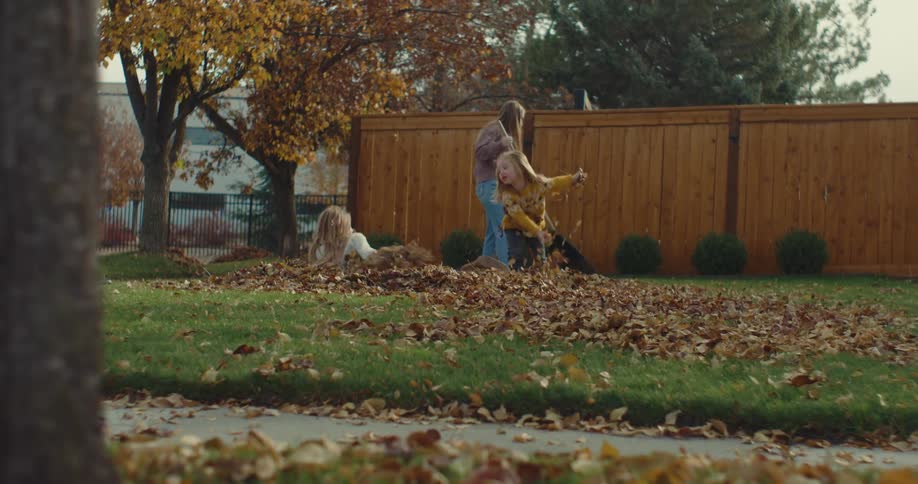 Adult using a rake to gather dry leaves in a backyard while two children play on a pile of leaves