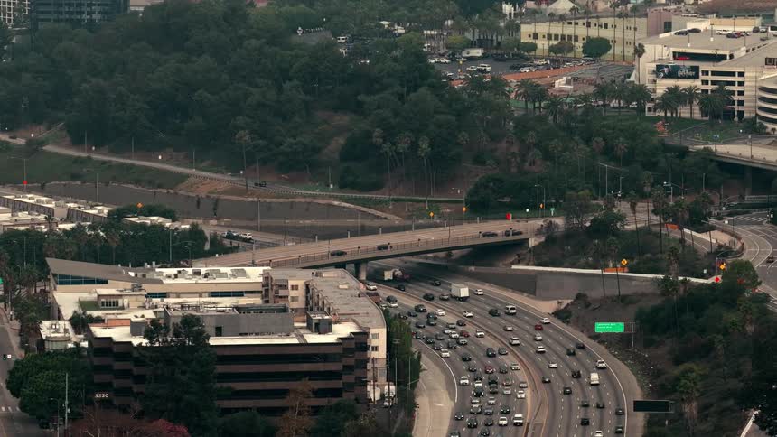 Highangle view showing steady vehicle traffic moving along a curved multilane freeway passing under a concrete overpass near hillside greenery apartment complexes and parking structures