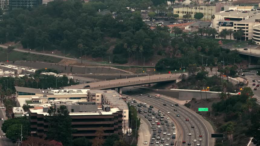 A high angle view shows steady traffic flowing along a multi lane highway as a black car crosses an overpass in a sprawling city under an overcast sky