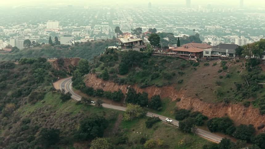 A distant view shows luxury houses perched on a green hillside above a winding road with a sprawling city visible in the background under a hazy sky