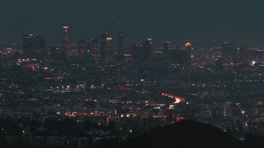 A distant view shows the illuminated skyline of los angeles at night with city lights and traffic stretching across the vast urban landscape under a dark sky