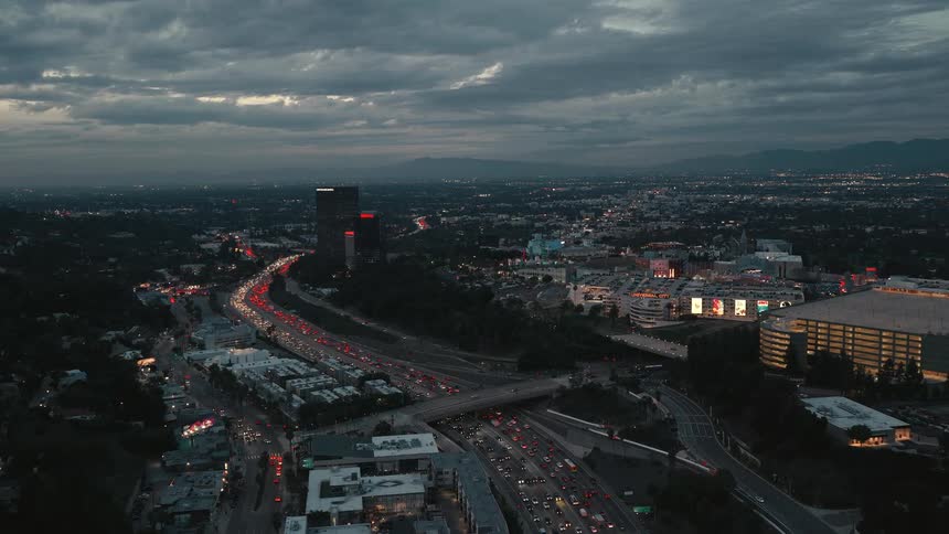 A wide aerial view shows heavy traffic flowing along a winding highway through a sprawling city at twilight under a dramatic cloudy sky
