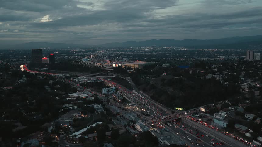 An aerial drone shot captures a vast city at dusk showing heavy traffic flow on a multilane highway winding through a valley of residential hills and commercial skyscrapers under a cloudy evening sky