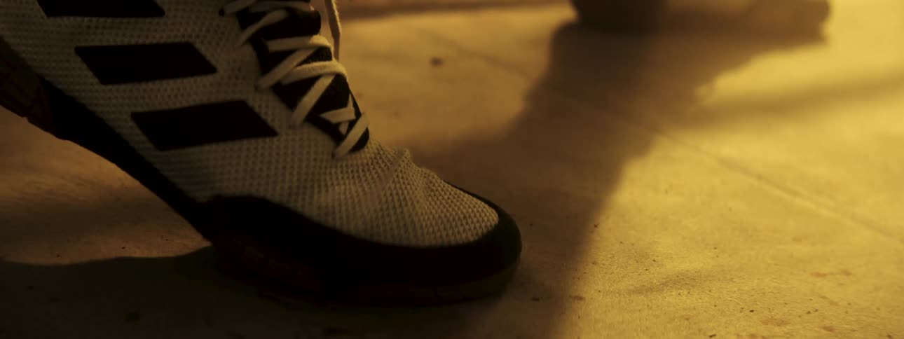 A close view shows the front of a white mesh boxing shoe with black stripes as spars in a dimly lit gym