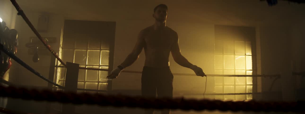 A man with a beard jumps rope inside a boxing ring in a dimly lit gym with light streaming through windows in the background