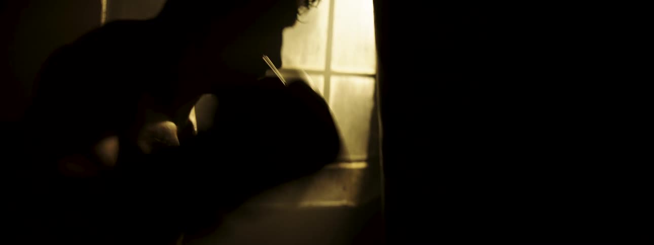A shirtless male boxer training in a dark gym striking a punching bag as warm bright light shines into the room