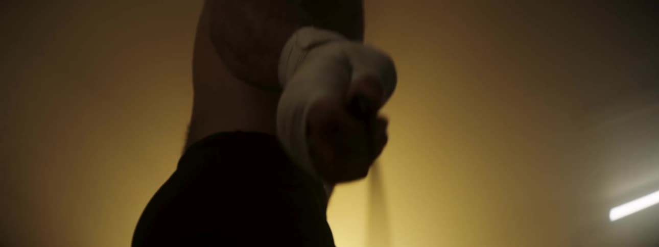 A close view shows the muscular torso of a shirtless man with wrapped hands jumping rope in a dimly lit gym with a warm orange glow