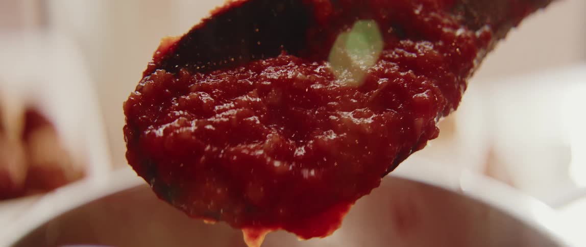 A close view shows thick red tomato sauce dripping from a wooden spoon held over a cooking pot