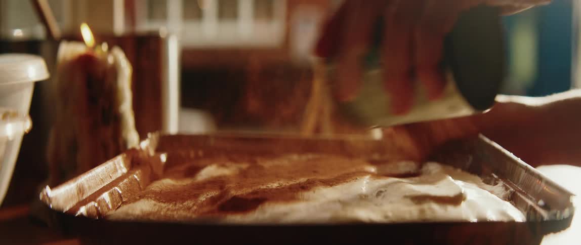 A person sprinkles a fine dark powder over a creamy dessert in a rectangular metal pan while lit candles flicker in the blurred background