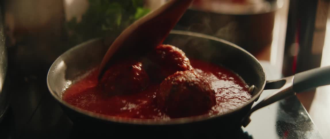 A wooden spoon bastes steaming meatballs as they simmer in a rich tomato sauce
