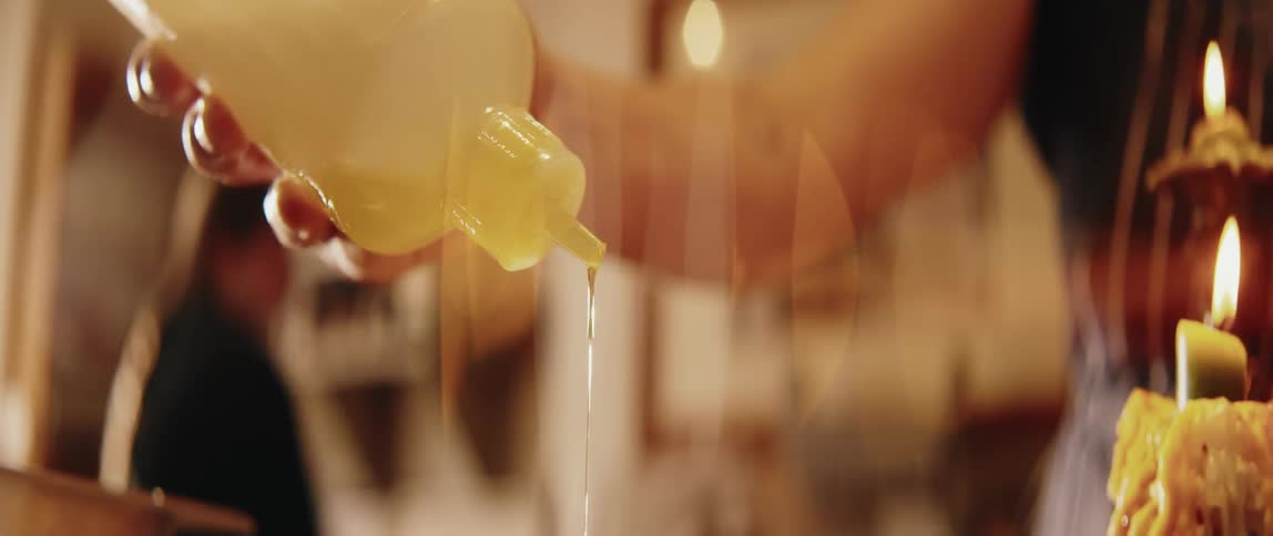A close view shows a stream of yellow oil being squeezed from a clear plastic bottle over a dish