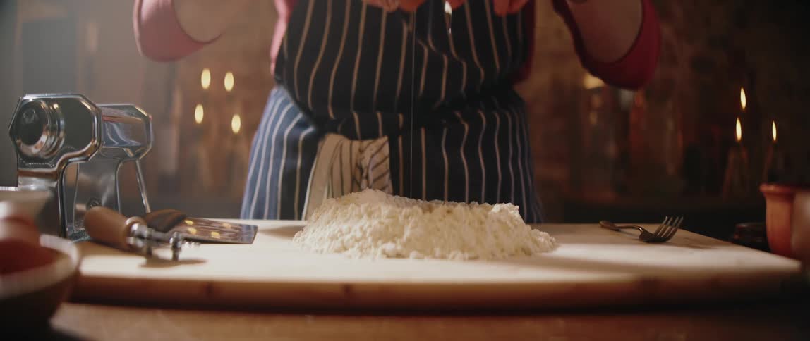 Person cracking an egg over a mound of flour on a wooden countertop for dough preparation