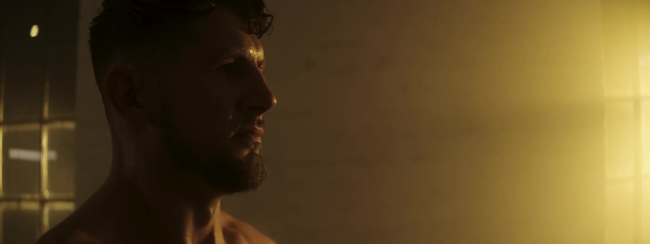 A shirtless sweaty man with a beard stands in a dimly lit room breathing heavily after a workout
